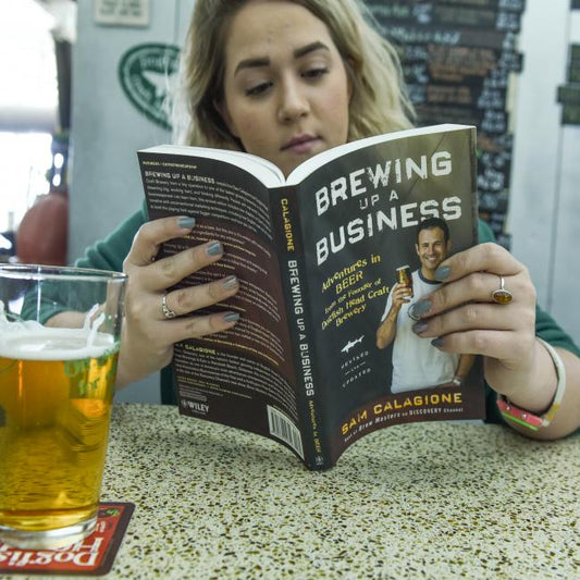 A woman sits at a speckled table reading "Book- Brewing Up A Busin" by J Wiley and Sons Publishing, with a glass of beer nearby—an inspiring scene for aspiring entrepreneurs.