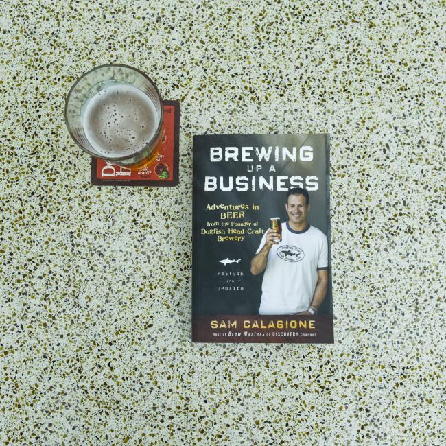 A glass of beer on a coaster sits beside the book "Brewing Up A Busin" by J Wiley and Sons Publishing on a speckled countertop. The book cover shows a man holding a bottle of beer.