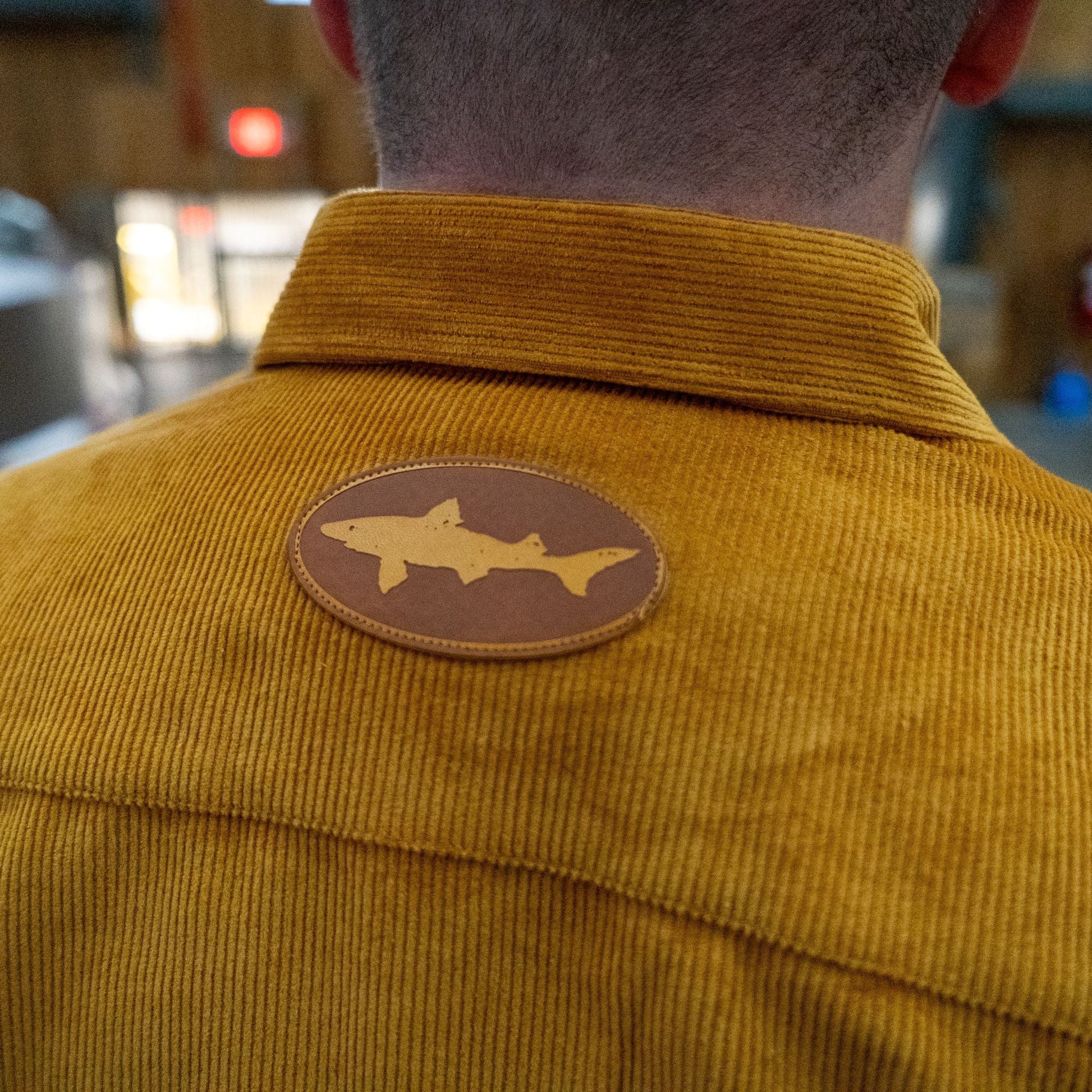 A person wears the Camel Cord Flannel Button Down by Brewery Branding, Inc., featuring camel-toned corduroy and a round brown shark silhouette patch on the upper back. The image highlights both the patch detail and textured fabric.