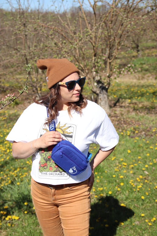 Front view of blue crisp belt bag on a woman. Angry Orchard bitten apple featured on bottom right of smaller pocket. Has writing "Angry Orchard" in white along strap.
