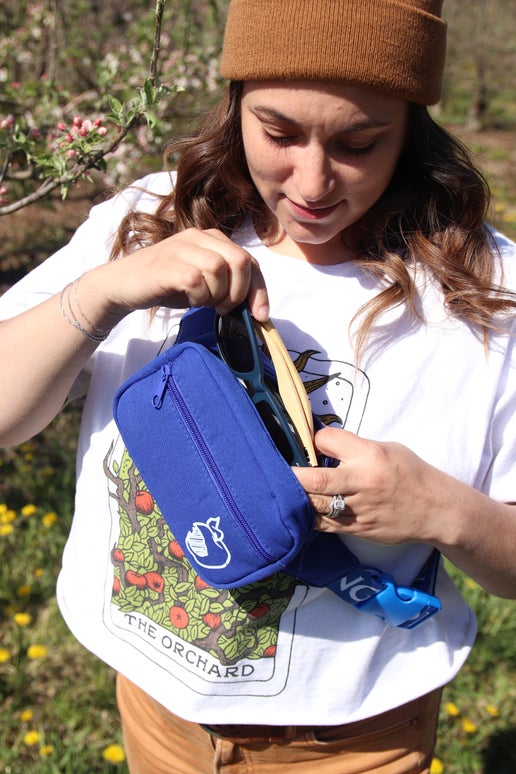Front view of blue crisp belt bag featuring a woman inserting an item into the bag. Angry Orchard bitten apple featured on bottom right of smaller pocket. Has writing "Angry Orchard" in white along strap.