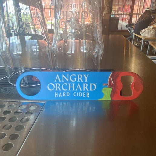 This blue paddle opener has the writing "Angry Orchard Hard Cider" in the center. On one end is a red apple which cuts open for the opener.