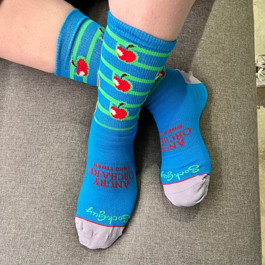 Blue mid-calf socks. Has writing "Angry Orchard Hard Cider" on foot and "Sock Guy" near toe area. Calf part of sock is striped green and has the red bitten Angry Orchard apples along it.