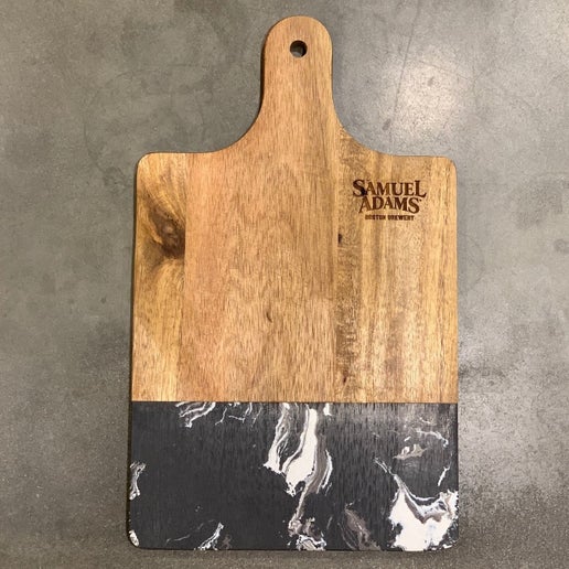 This is a rectangular cutting wood cutting board, with a black marble design on the bottom half of the wood and a Samuel Adams logo on the top right corner