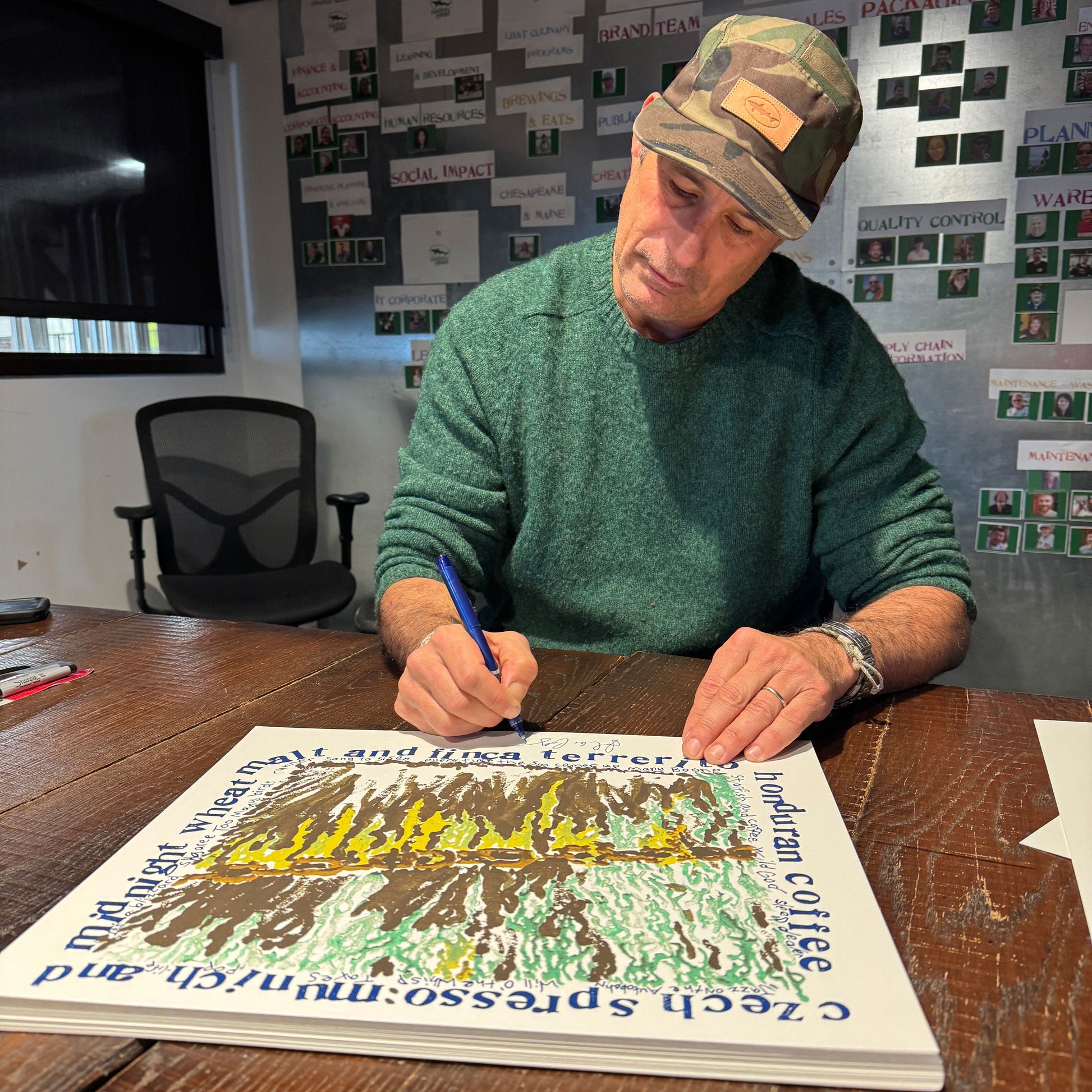 Man signing a poster at a desk with a wall of posters in the background