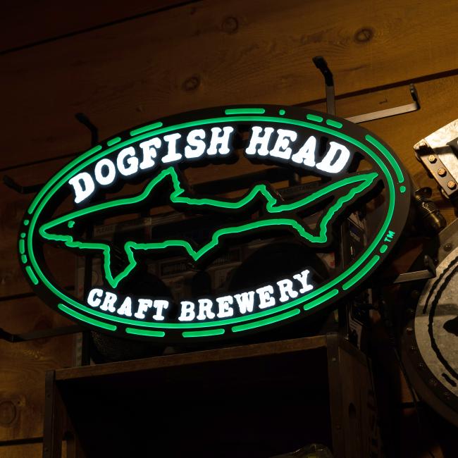 The Merchtank Craft Brewery Logo LED Neon features an oval design with a shark and “Dogfish Head Craft Brewery” in white and green, mounted on a wooden wall.