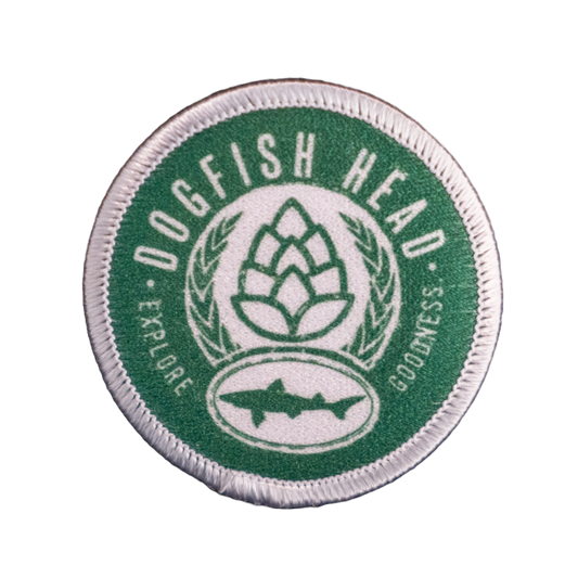 The Explore Goodness Patch by GRAFFITI INC is a circular green and white patch featuring “Dogfish Head” at the top, a hop cone with laurel leaves in the center, a dogfish silhouette at the bottom, plus “every sip,” “Explore,” and “Sessions.”.