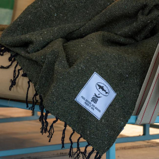 A dark green DFH INN Blanket by Trek Light Gear with black fringes is draped over a beige cushion. Made from recycled materials, it has a white patch in the corner with a fish logo and ‘FEEL GOOD. DO GOOD. PASS IT ON.’.