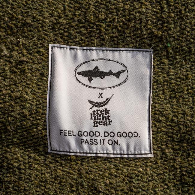 A white fabric patch on green DFH INN Blanket material displays a fish logo above "FEEL GOOD. DO GOOD. PASS IT ON." and the Trek Light Gear brand logo, highlighting its eco-friendly upcycled design.