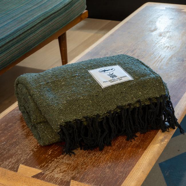 The DFH INN Blanket by Trek Light Gear, a handmade dark green piece with black fringes and a white patch, is folded on a wooden bench. A striped cushion is partially visible in the background.