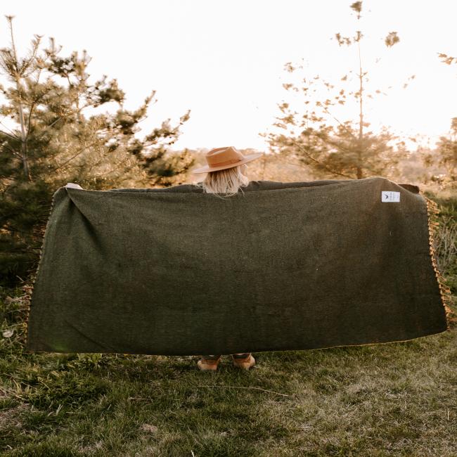 A person standing on grass with their back to the camera holds a Trek Light Gear DFH INN Blanket spread wide, wearing a brown hat. Sunlit pine trees fill the outdoor background.