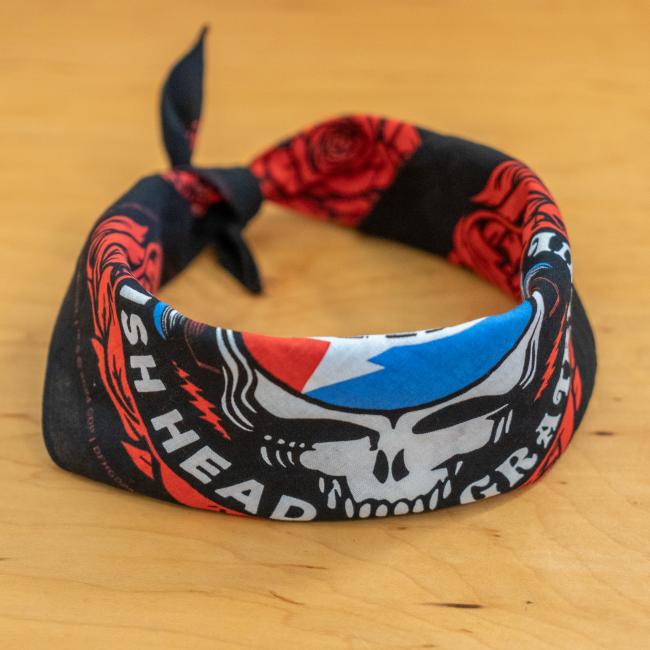 The Dogfish Head x Grateful Dead Black Bandana by Merchtank, featuring red roses and a white skull with a blue and red lightning bolt, sits on a light wood surface. The text "HEAD GR" adds to its unique craft beer accessory appeal.