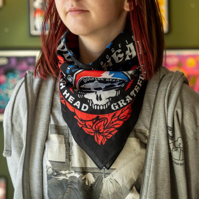A person with red-streaked hair wears a Merchtank Dogfish Head x Grateful Dead Black Bandana featuring a skull design over a gray graphic T-shirt and light gray hoodie. Colorful posters appear in the blurred background.