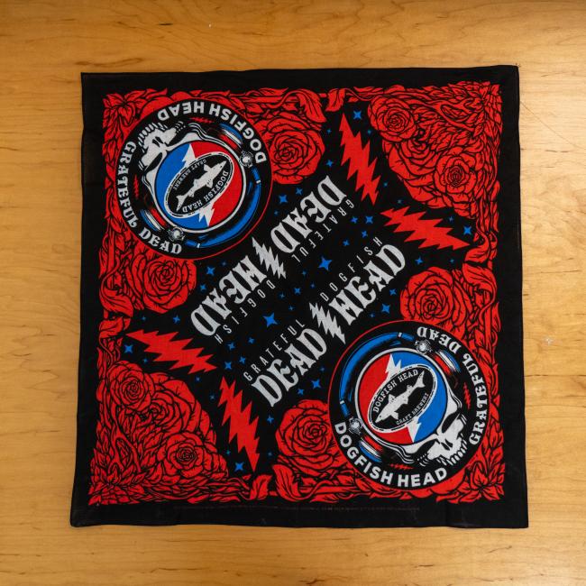 The Merchtank Dogfish Head x Grateful Dead Black Bandana features Dogfish Head logos, red roses, and lightning bolts on a black background with blue accents—a bold craft beer accessory displayed on wood.