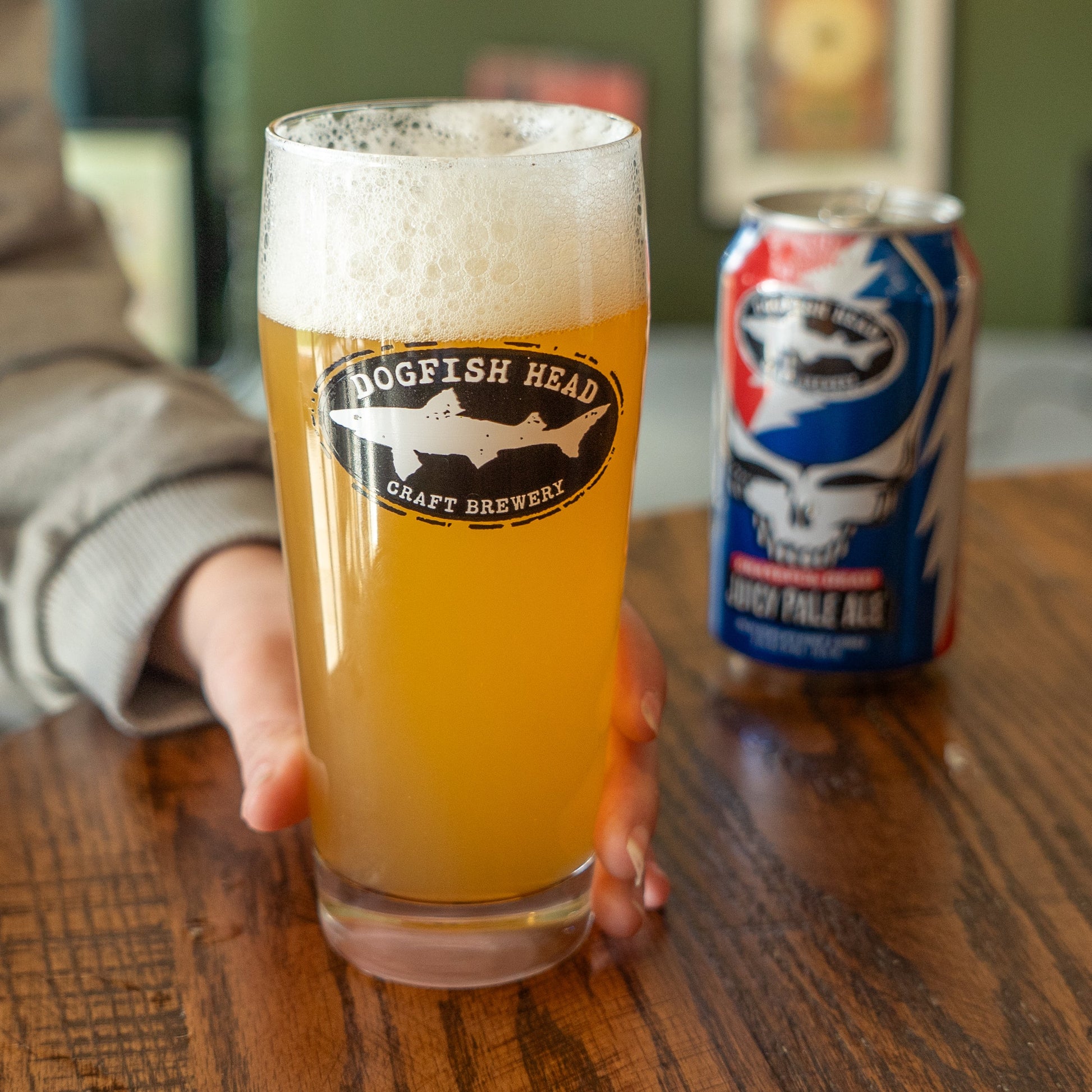 A person holds a Dogfish Head x Grateful Dead Juicy Glass by Grandstand Glassware & Apparel, filled with hazy craft beer on a wooden table. A colorful can and Grateful Dead merchandise appear blurred in the background, enhancing the vibe.