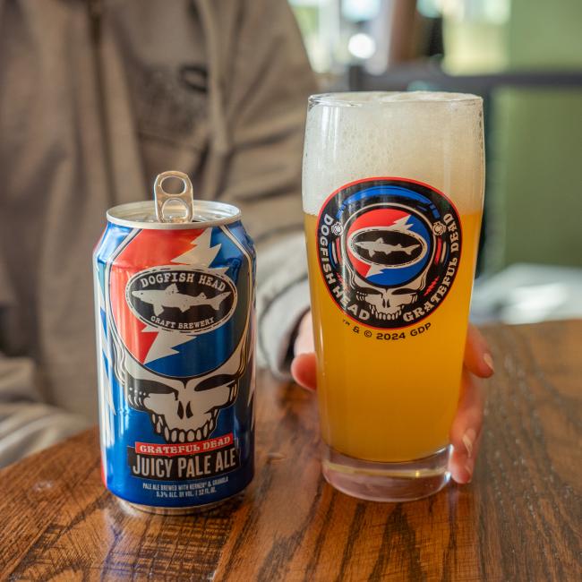 A person holds a Dogfish Head x Grateful Dead Juicy Glass by Grandstand Glassware & Apparel beside an open can of Dogfish Head Grateful Dead Juicy Pale Ale on a wooden table—ideal for craft beer and Grateful Dead fans.