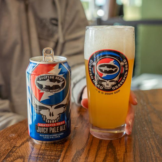 A person holds a Dogfish Head x Grateful Dead Juicy Glass by Grandstand Glassware & Apparel beside an open can of Dogfish Head Grateful Dead Juicy Pale Ale on a wooden table—ideal for craft beer and Grateful Dead fans.