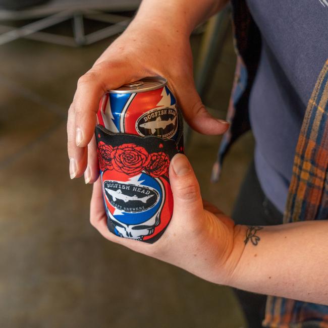 A person holds a Dogfish Head beer in a Dogfish Head x Grateful Dead Skull and Roses Can Cooler by Grandstand Glassware & Apparel, showing off the red roses and blue skull. A wrist tattoo and plaid shirt add to the relaxed festival vibe.