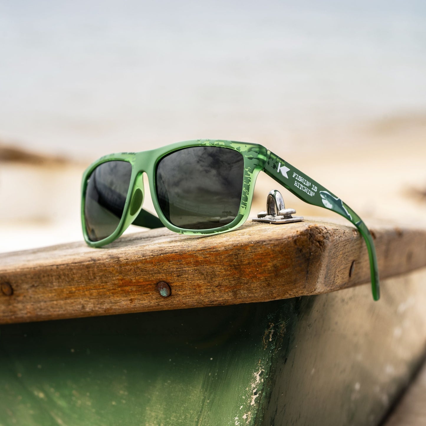 Green sunglasses with dark lenses resting on the edge of a weathered wooden boat, with a blurred beach background.