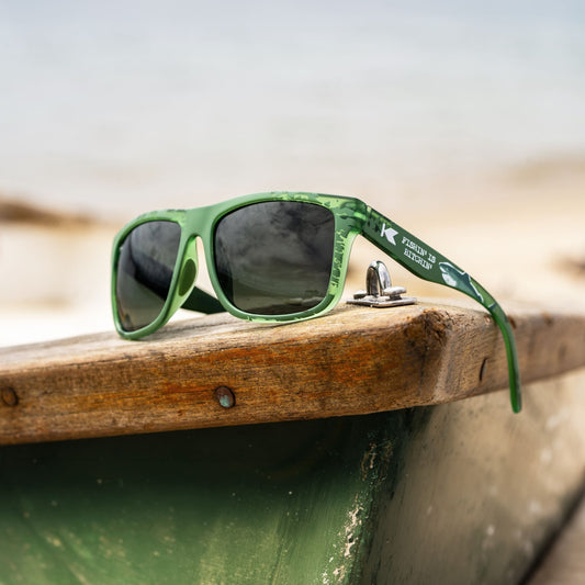 Green sunglasses with dark lenses resting on the edge of a weathered wooden boat, with a blurred beach background.