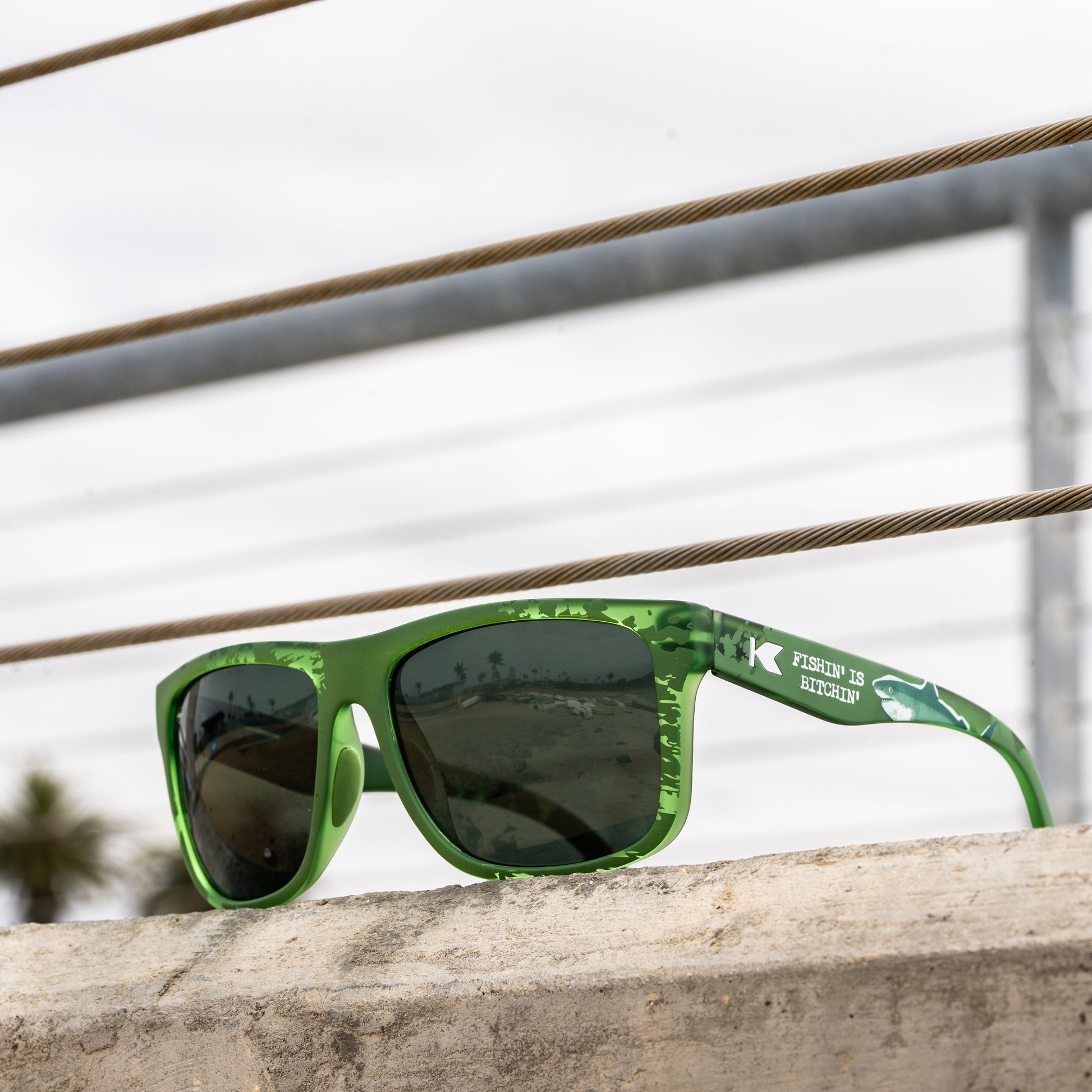 Green sunglasses with a subtle camouflage pattern on the frames and dark lenses, resting on a concrete ledge outdoors.