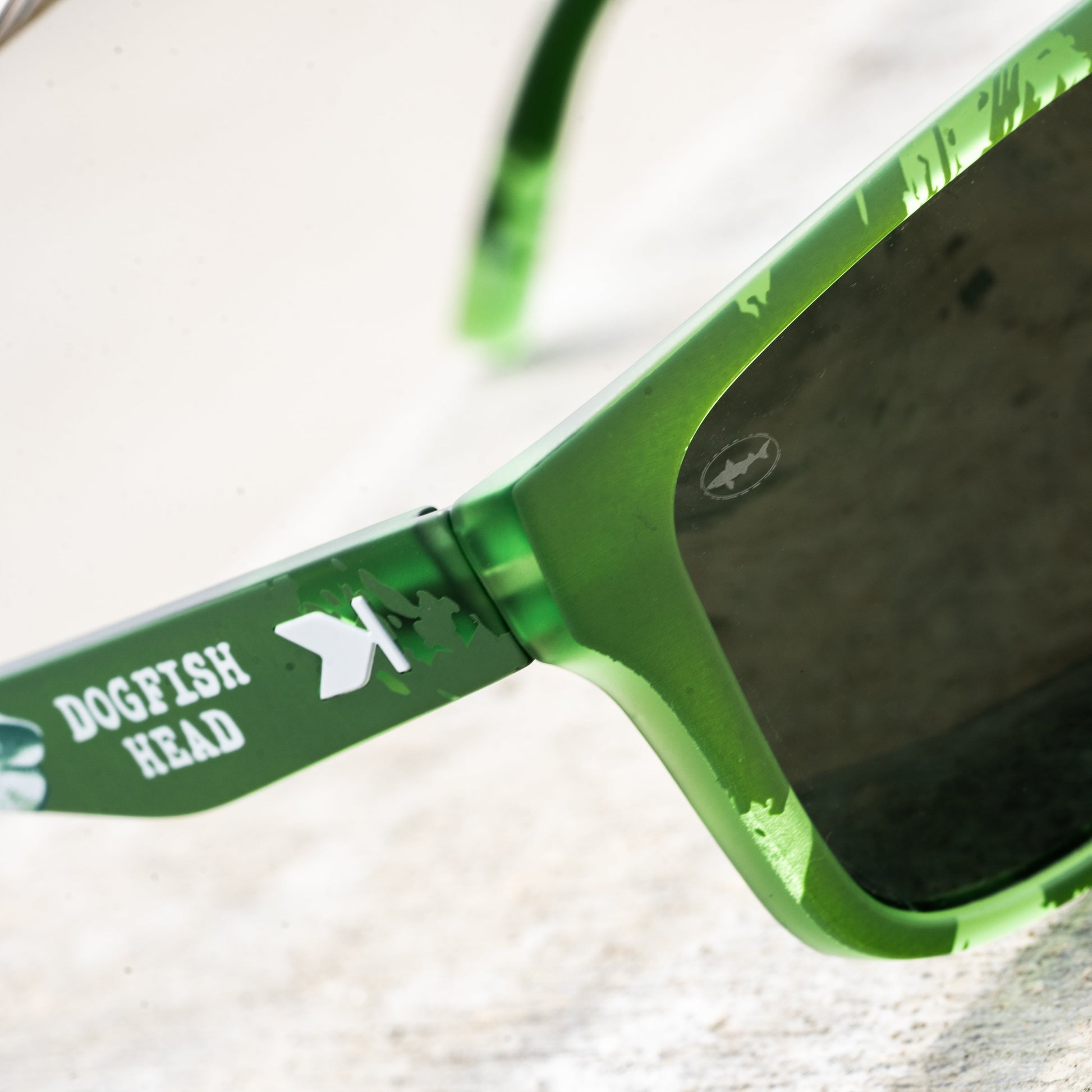 Close-up of green sunglasses with dark lenses and a green temple arm printed with the text 'DOGFISH HEAD' and a white fish logo, resting on a light textured surface.