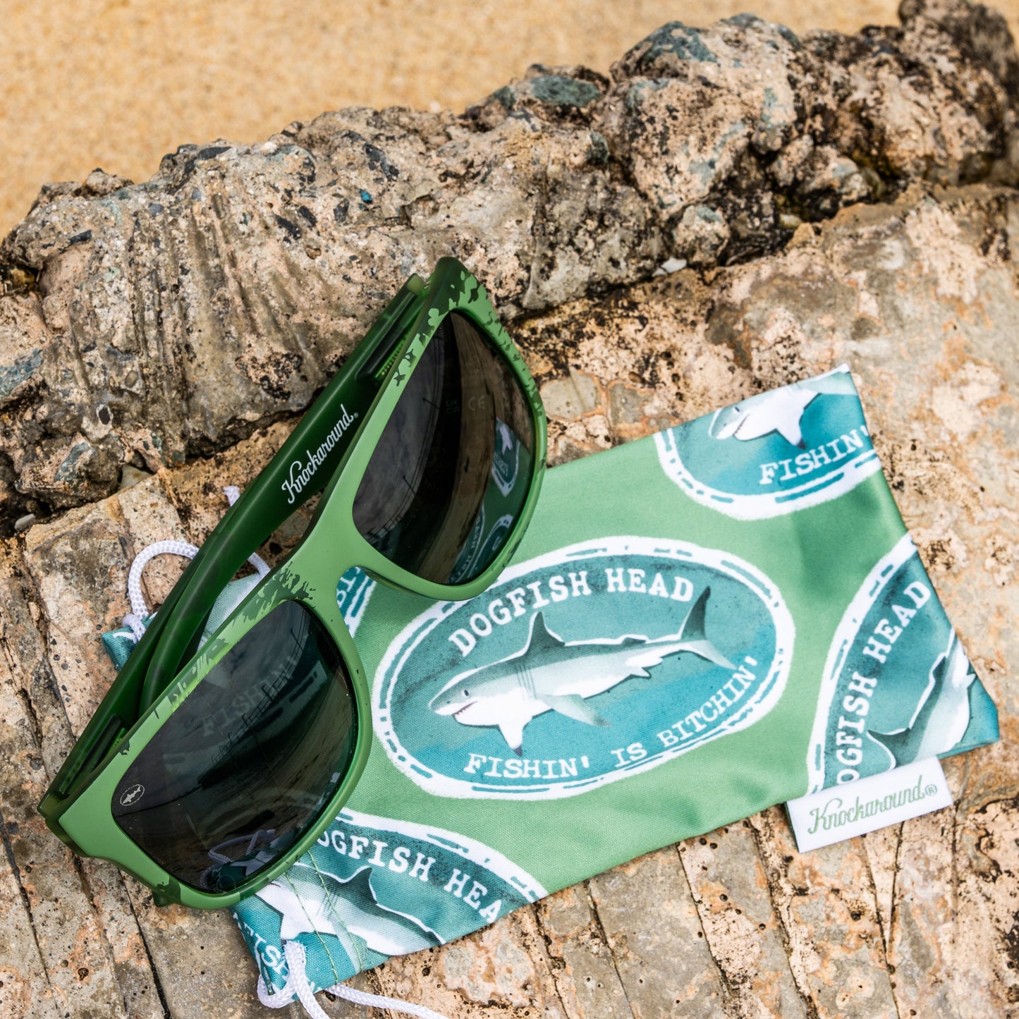 Green Dogfish Head sunglasses resting on a green branded bag, placed on a textured rock with sand in the background.