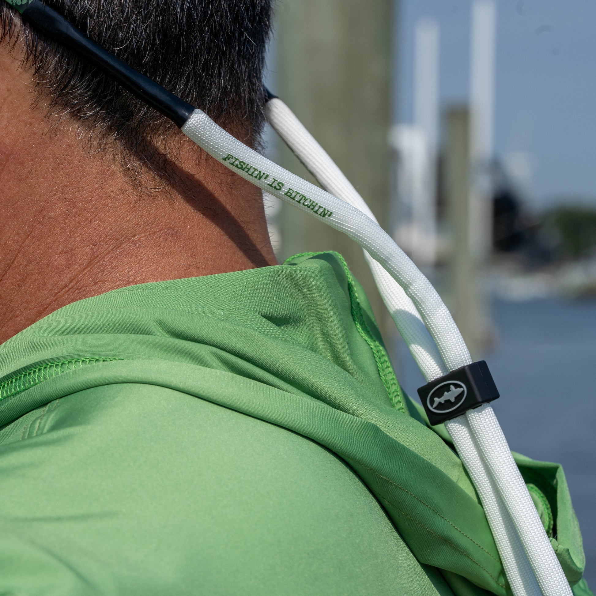 Close-up of a person wearing a green hooded tee outdoors, with white drawstrings attached to sunglasses with shark logo adjustor. Sunglasses are also visible on the head.