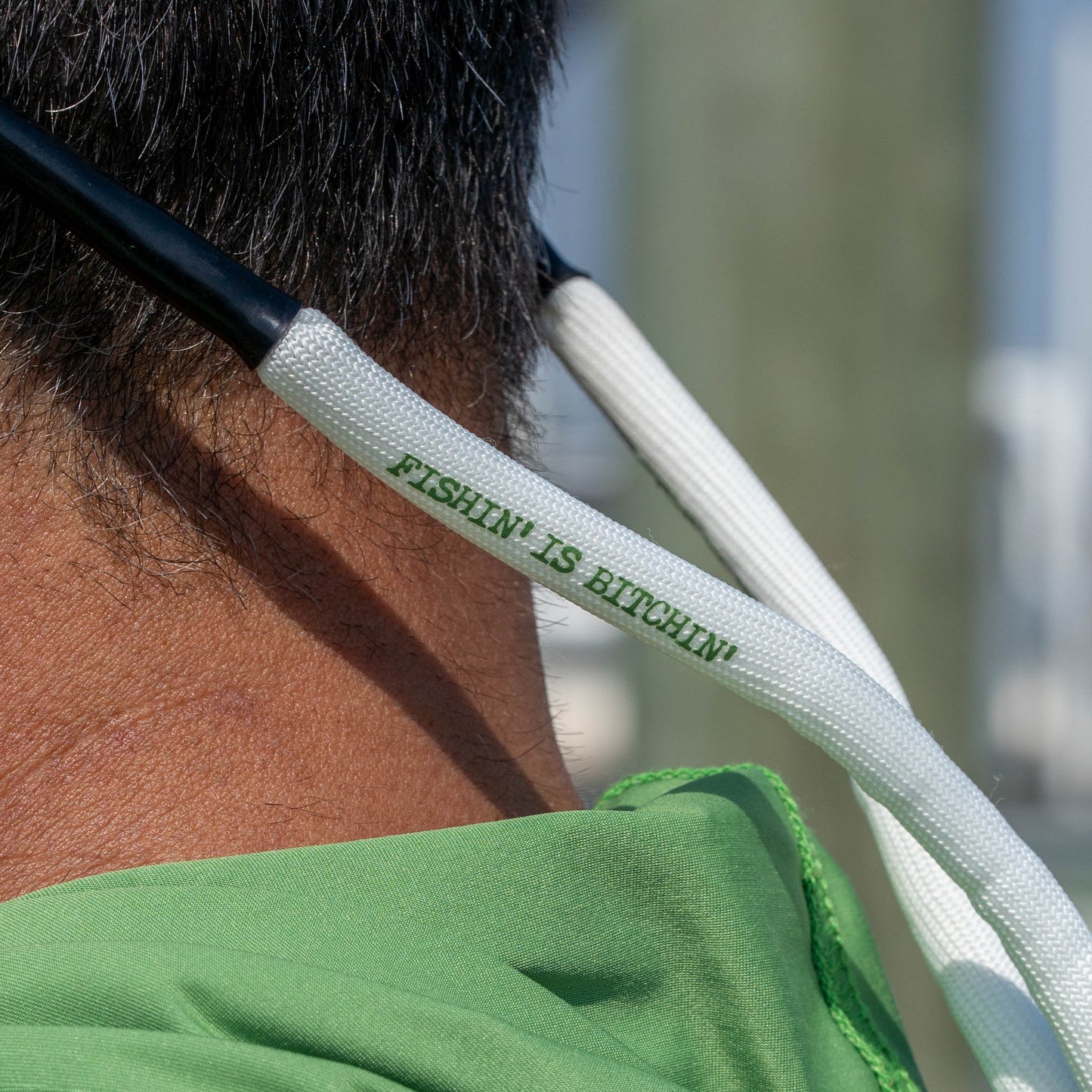 Close-up of a person outdoors wearing a white woven neck strap with green text reading 'FISHIN' IS BITCHIN''.