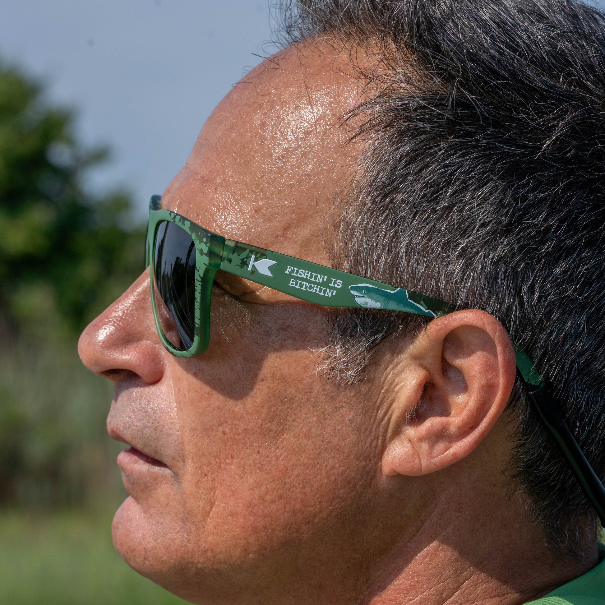 Close-up side profile of a man wearing green sunglasses outdoors. The sunglasses have dark lenses and a green frame with white text reading 'FISHING IS BITCHLIN' and a shark graphic on the temple.