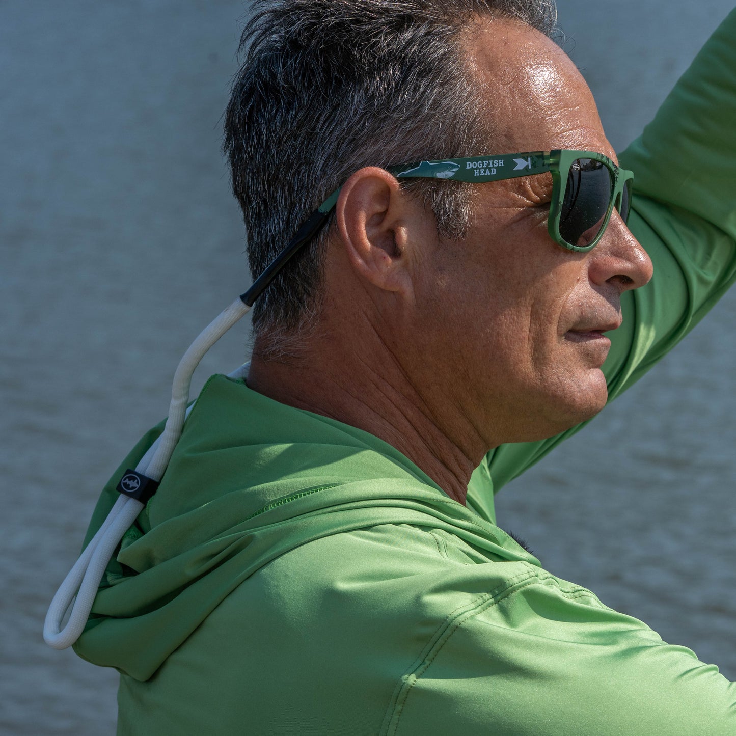 A man in a green hooded shirt and green-framed sunglasses with a white neck cord, photographed outdoors against a blurred water background.