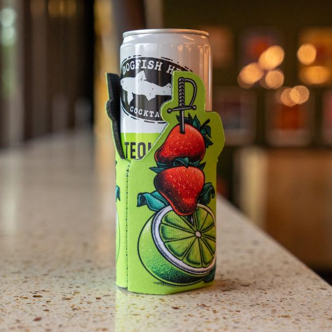 A Merchtank Four Slim Can Cooler Bundle holds a white 12oz Dogfish Head can in a green koozie with sliced lime, strawberries, and cocktail pick illustrations, set on a countertop with blurred lights in the background.