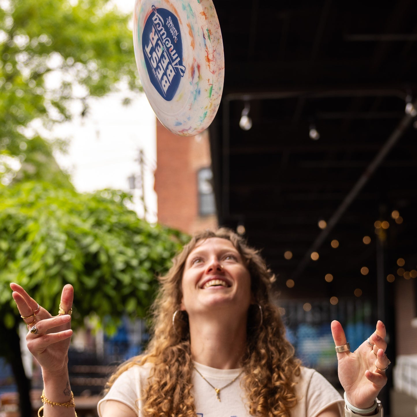 A person throwing a frisbee with a white base but some multicolor accents, featuring a navy shield on the front with the phrase "The best beer in America" in it. Also features the Sam Adams logo.