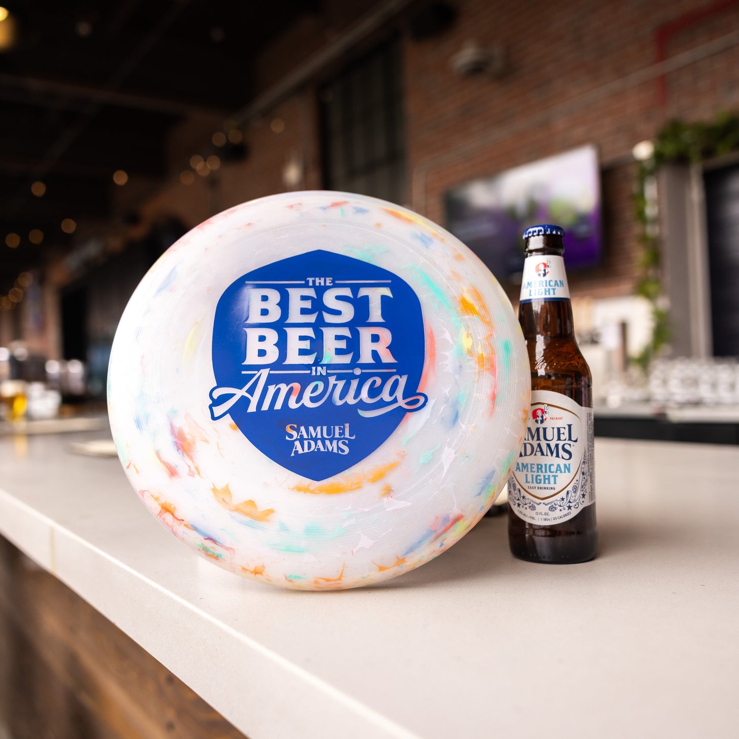 A frisbee with a white base but some multicolor accents, featuring a navy shield on the front with the phrase "The best beer in America" in it. Also features the Sam Adams logo.