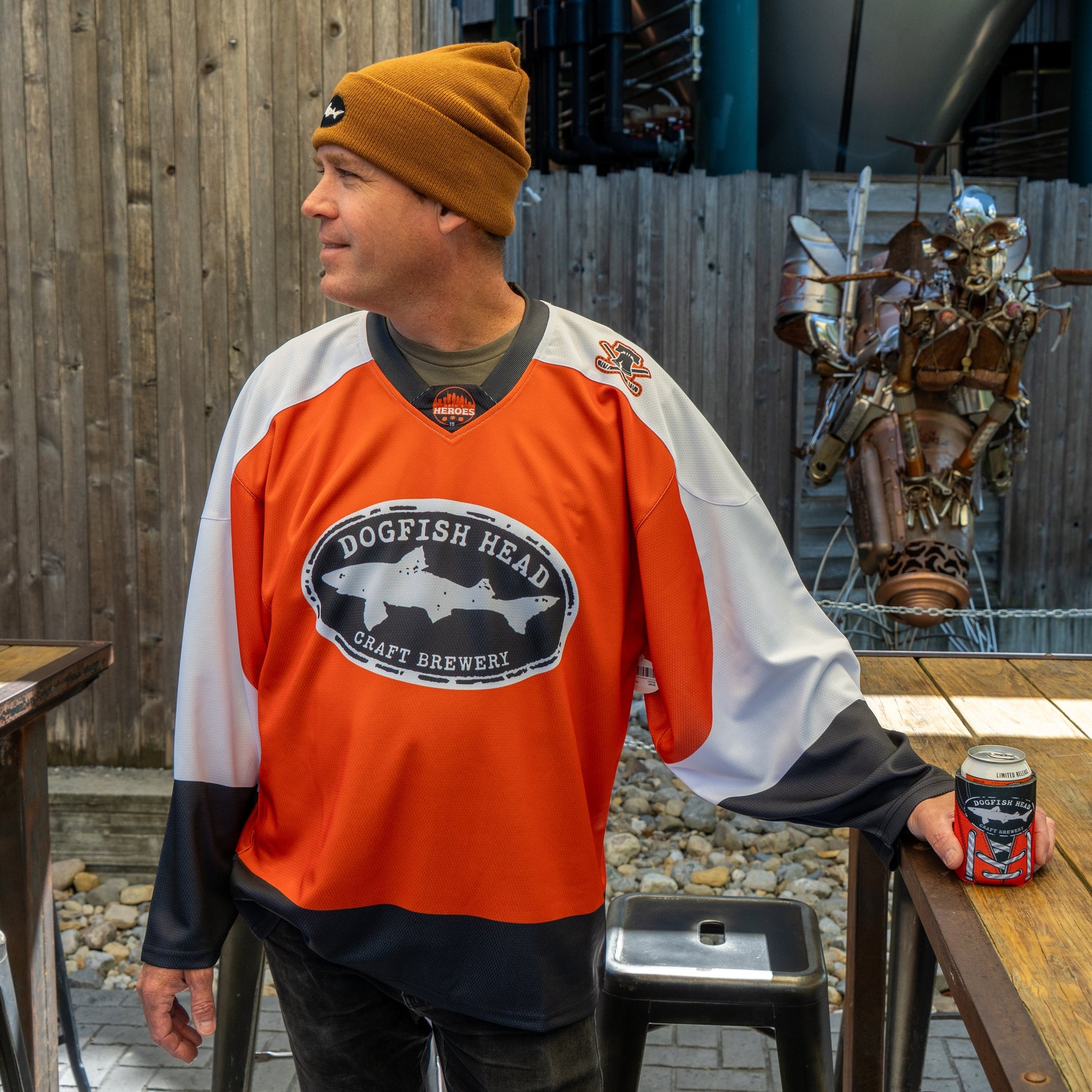 A man in an orange Dogfish Head x Garnet Hathaway Hockey Jersey by In Your Face Apparel and a brown beanie stands outdoors by a wooden table, holding a can. He looks to the side, with a metal sculpture and wooden fence behind him.