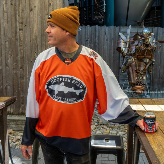 A man in an orange Dogfish Head x Garnet Hathaway Hockey Jersey by In Your Face Apparel and a brown beanie stands outdoors by a wooden table, holding a can. He looks to the side, with a metal sculpture and wooden fence behind him.