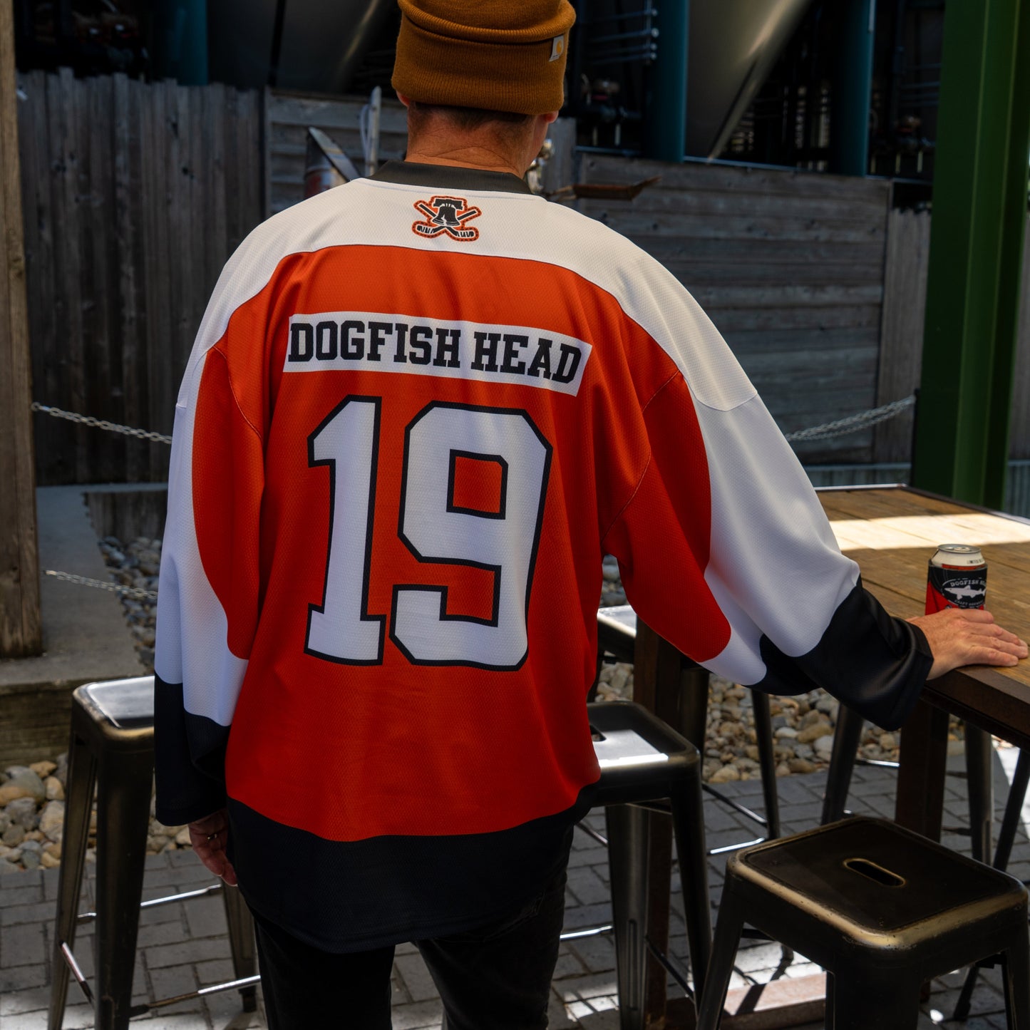 Someone outdoors wears an In Your Face Apparel Dogfish Head x Garnet Hathaway Hockey Jersey, holding a can and resting a hand on a metal chair; their face isn't visible.