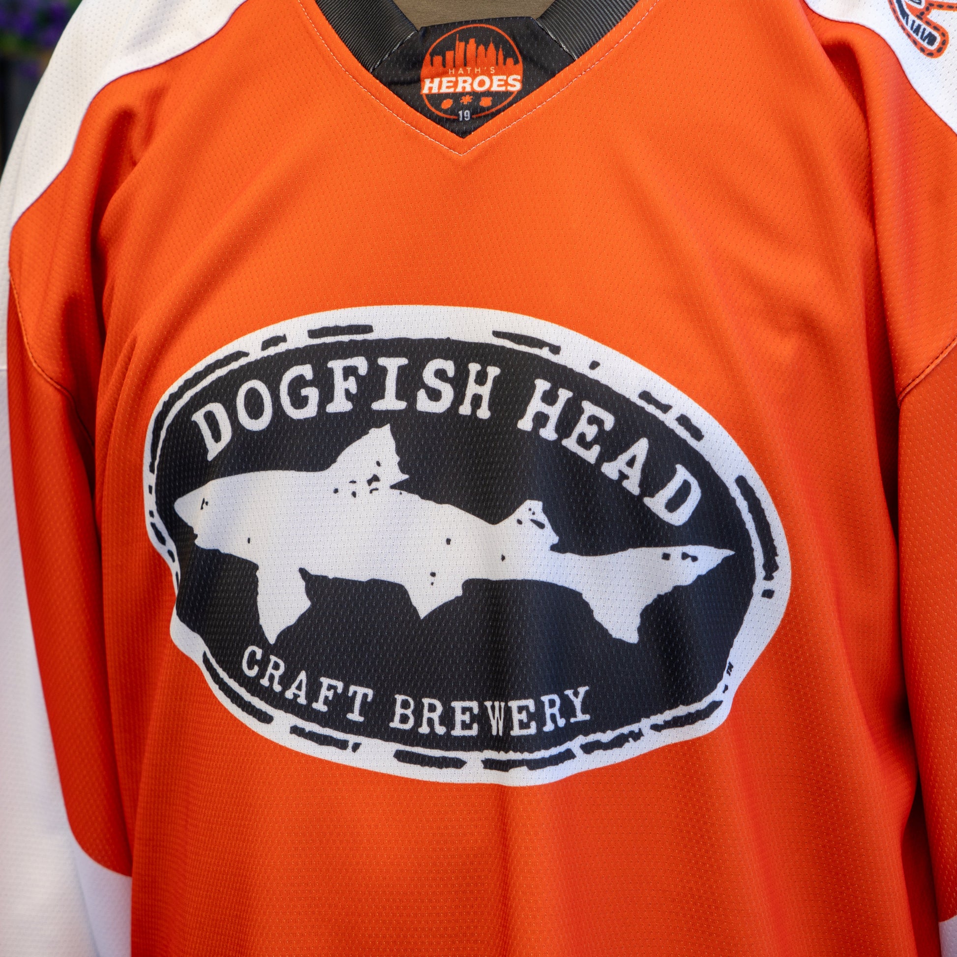 A close-up of the Dogfish Head x Garnet Hathaway Hockey Jersey by In Your Face Apparel, showcasing a black-and-white fish logo on the chest and a Heroes collar label that honors first responders.