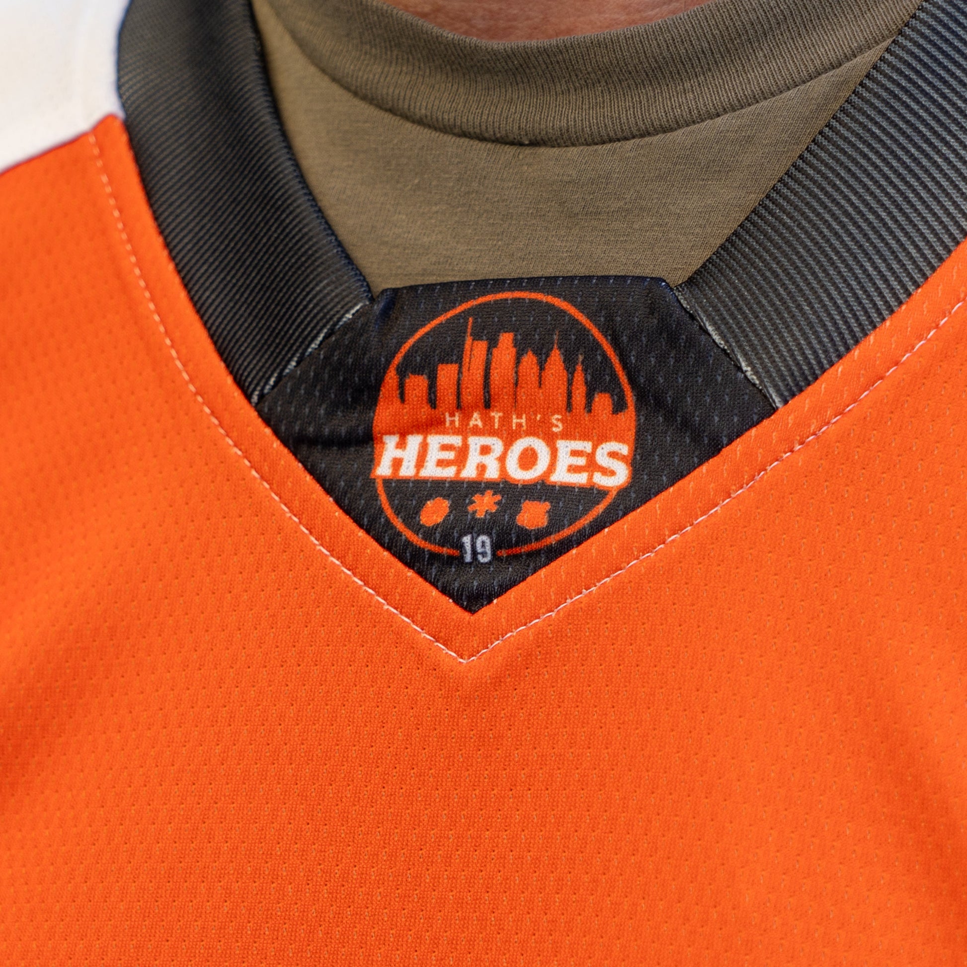 Close-up of the collar on an orange Dogfish Head x Garnet Hathaway Hockey Jersey by In Your Face Apparel, featuring a black patch with "Haths Heroes," a city skyline, icons, and number 19 honoring first responders.
