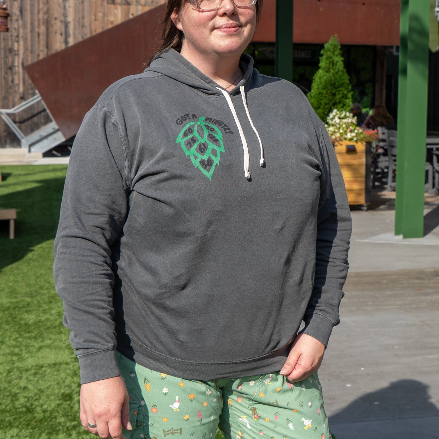 A person wearing glasses, a Grey Got A Minute Hoodie by GO WEST T-SHIRT COMPANY and light green patterned pajama pants stands outdoors on a sunny day in front of a wooden building, their face partly visible.