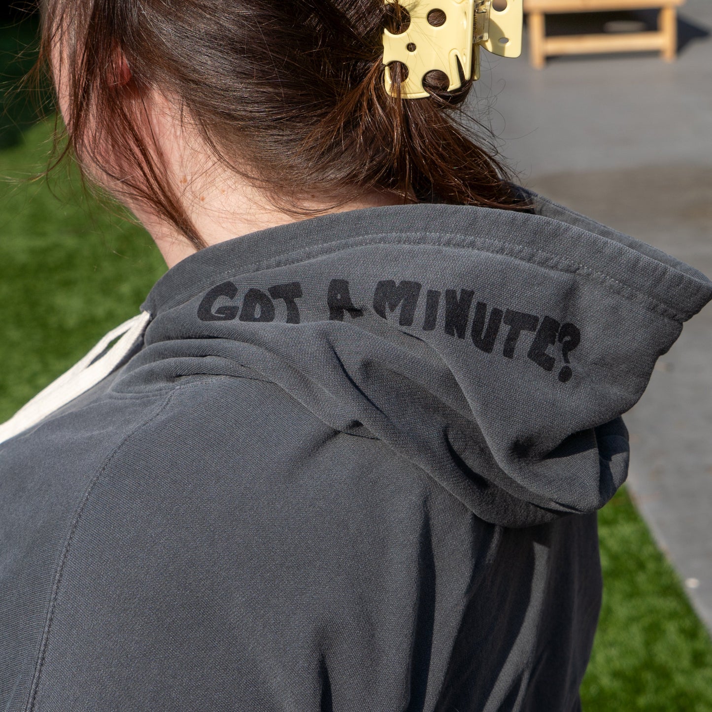 Someone wears the GO WEST T-SHIRT COMPANY’s Grey Got A Minute Hoodie, featuring "GOT A MINUTE?" on the hood. Their brown hair is tied back with a yellow clip, and grass with pavement can be seen outdoors in the background.