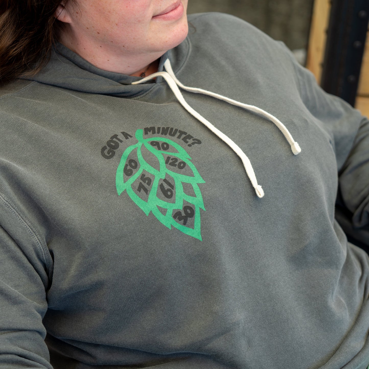 A person wears the Grey Got A Minute Hoodie by GO WEST T-SHIRT COMPANY, featuring a hop cone with clock numbers and "Got a minute?" text. This Minute Series beer apparel includes white drawstrings.