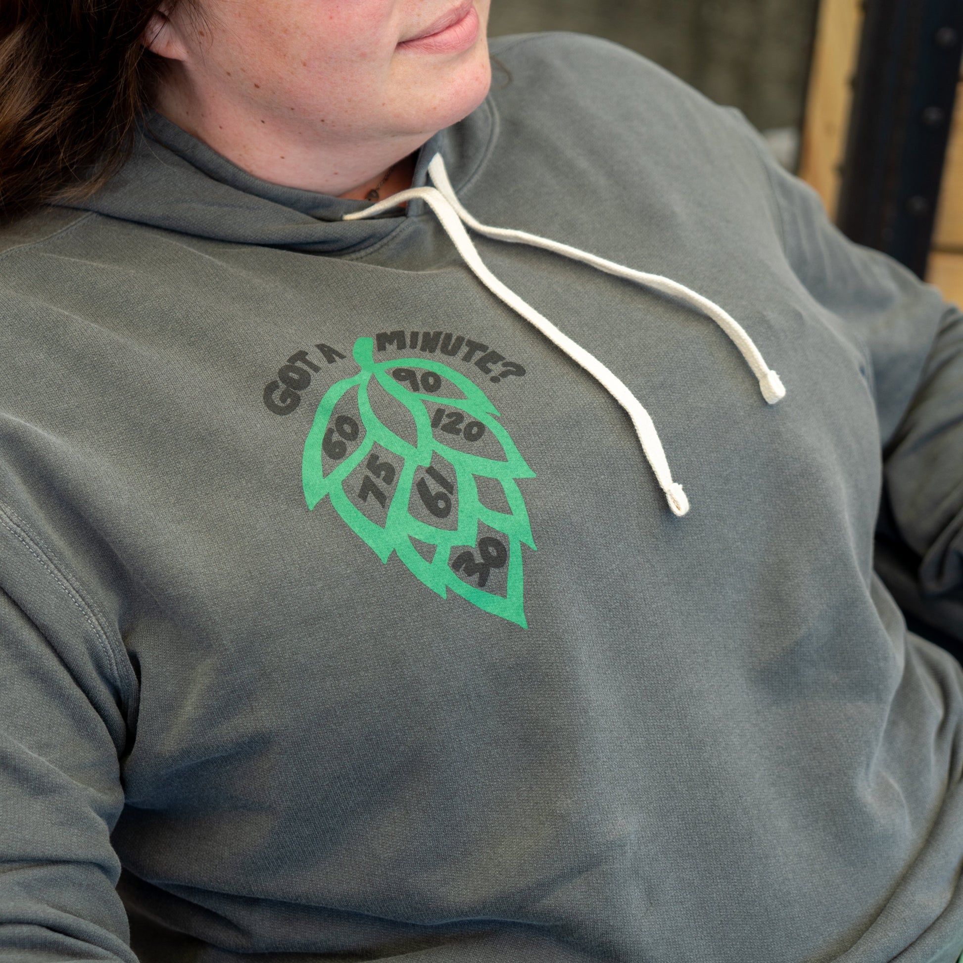 A person wears the Grey Got A Minute Hoodie by GO WEST T-SHIRT COMPANY, featuring a hop cone with clock numbers and "Got a minute?" text. This Minute Series beer apparel includes white drawstrings.
