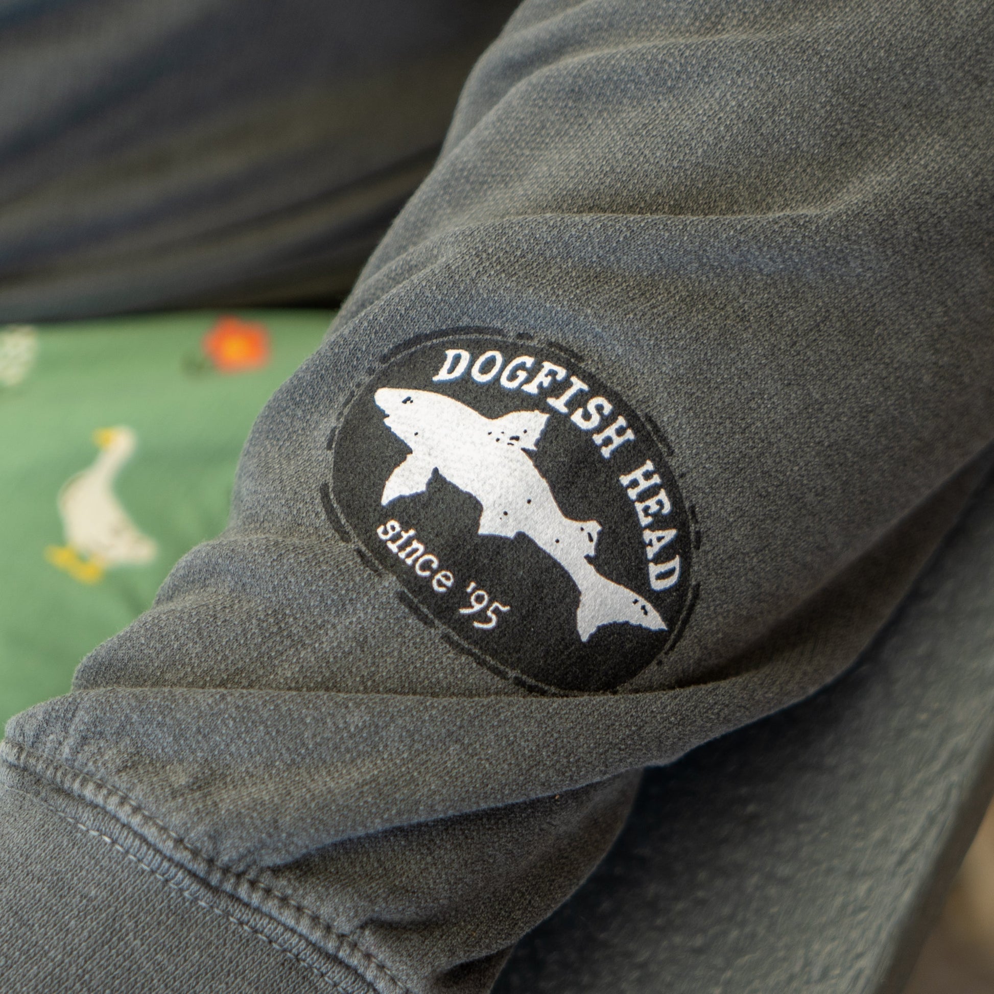 A close-up of the Grey Got A Minute Hoodie by GO WEST T-SHIRT COMPANY shows its sleeve with a Dogfish Head since 95 patch. In the background, green fabric with duck or goose illustrations appears. Perfect for beer apparel fans.