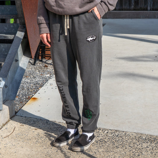 Outdoors, only the lower half is visible: a person wears GO WEST T-SHIRT COMPANY’s Grey Got A Minute Sweatpants featuring text and graphics on the left leg, gray sweatshirt, and black slip-on shoes.