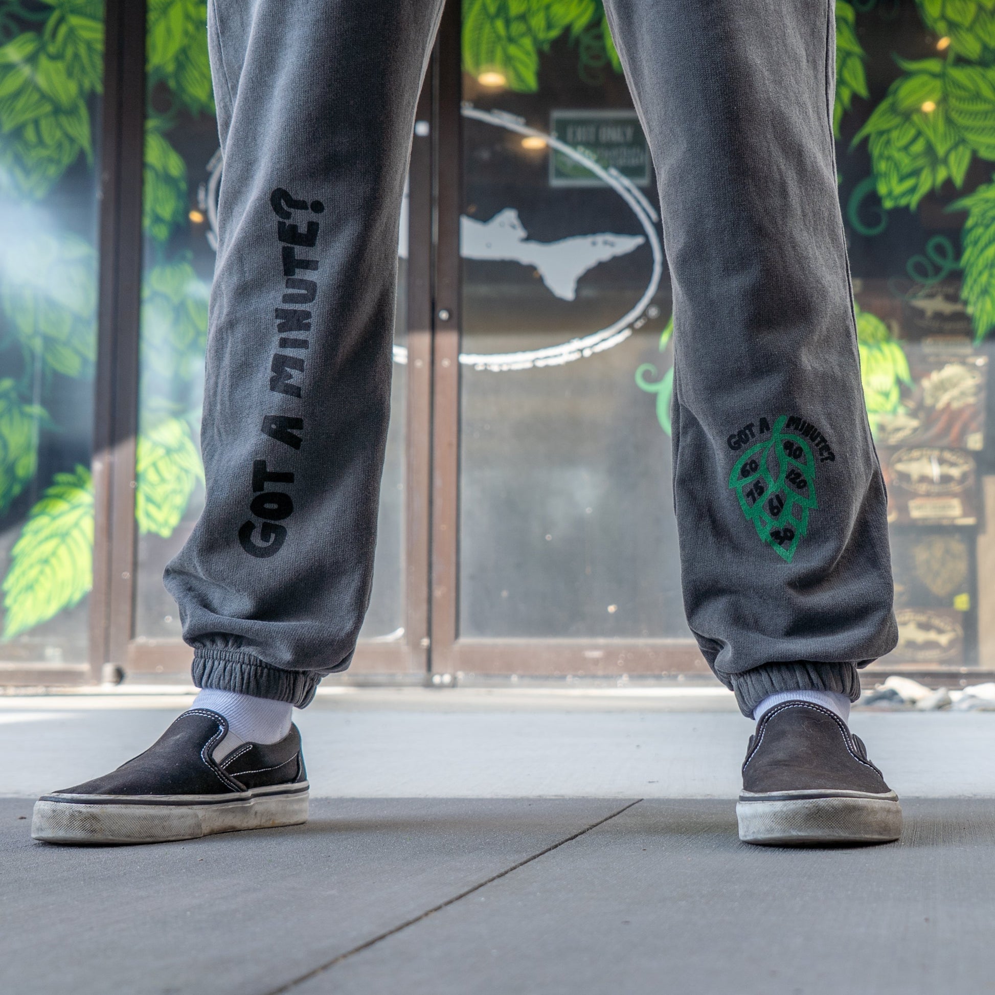 Person stands outside in GO WEST T-SHIRT COMPANY's Grey Got A Minute Sweatpants featuring "GOT A MINUTE?" and a green leaf graphic, paired with white socks and black slip-on shoes. Glass door and leafy mural set a relaxed vibe.
