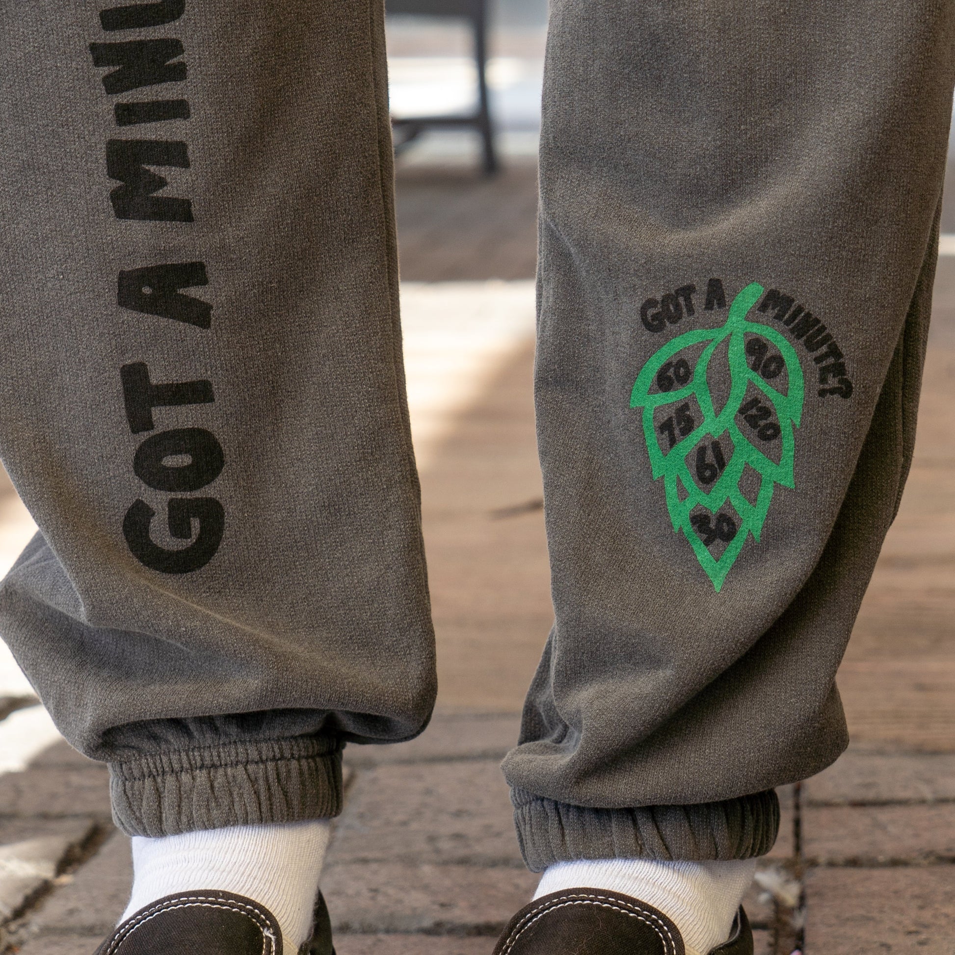Close-up of GO WEST T-SHIRT COMPANY’s Grey Got A Minute Sweatpants, detailed with “GOT A MINUTE?” and a green leaf graphic with numbers. Styled casually with white socks and dark shoes for easygoing comfort.
