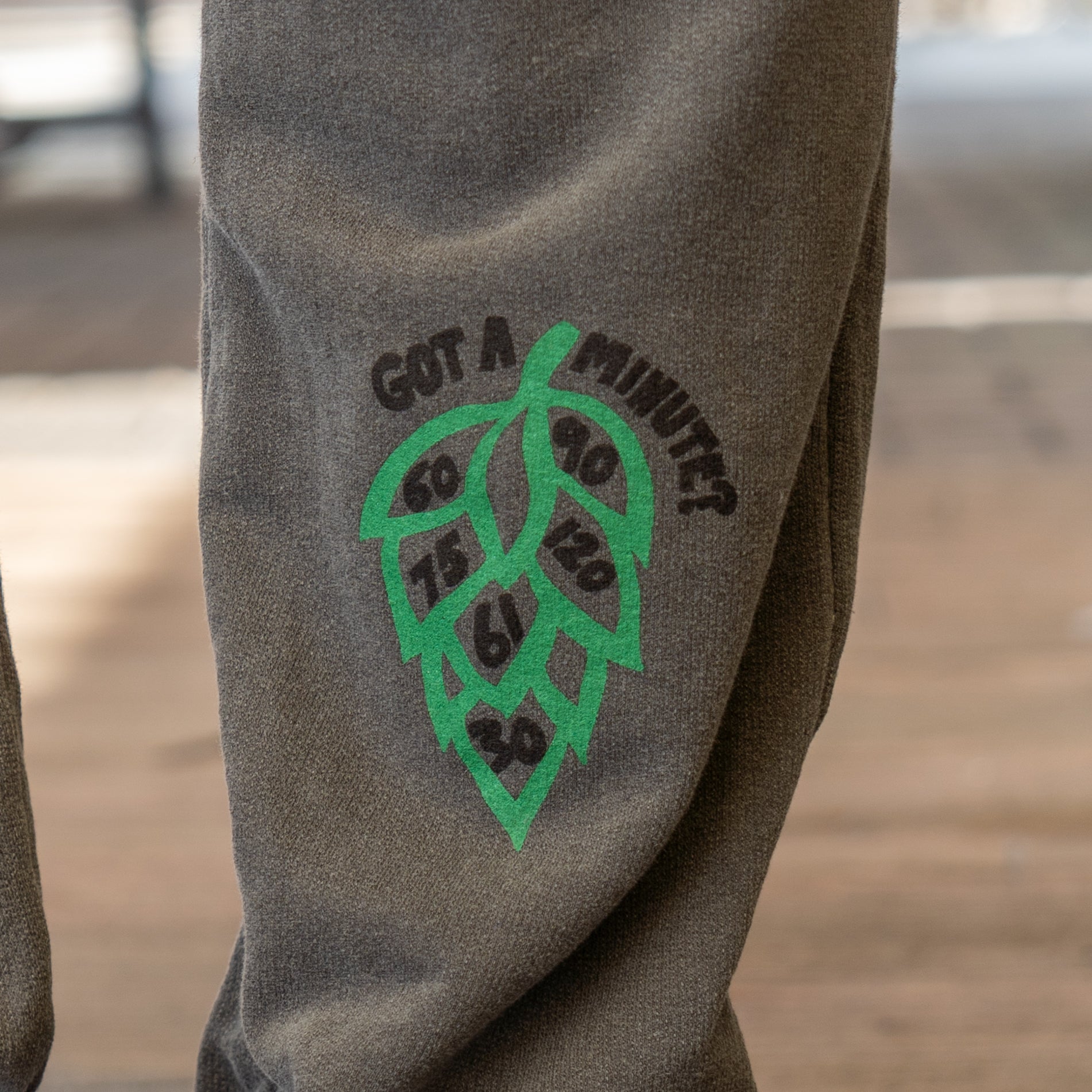 Close-up of GO WEST T-SHIRT COMPANY’s Grey Got A Minute Sweatpants shows a green hop cone with numbers 60, 90, 120, 75, 6, and 30 inside each leaf; "GOT A MINUTE?" is arced above the illustration on the pant leg.
