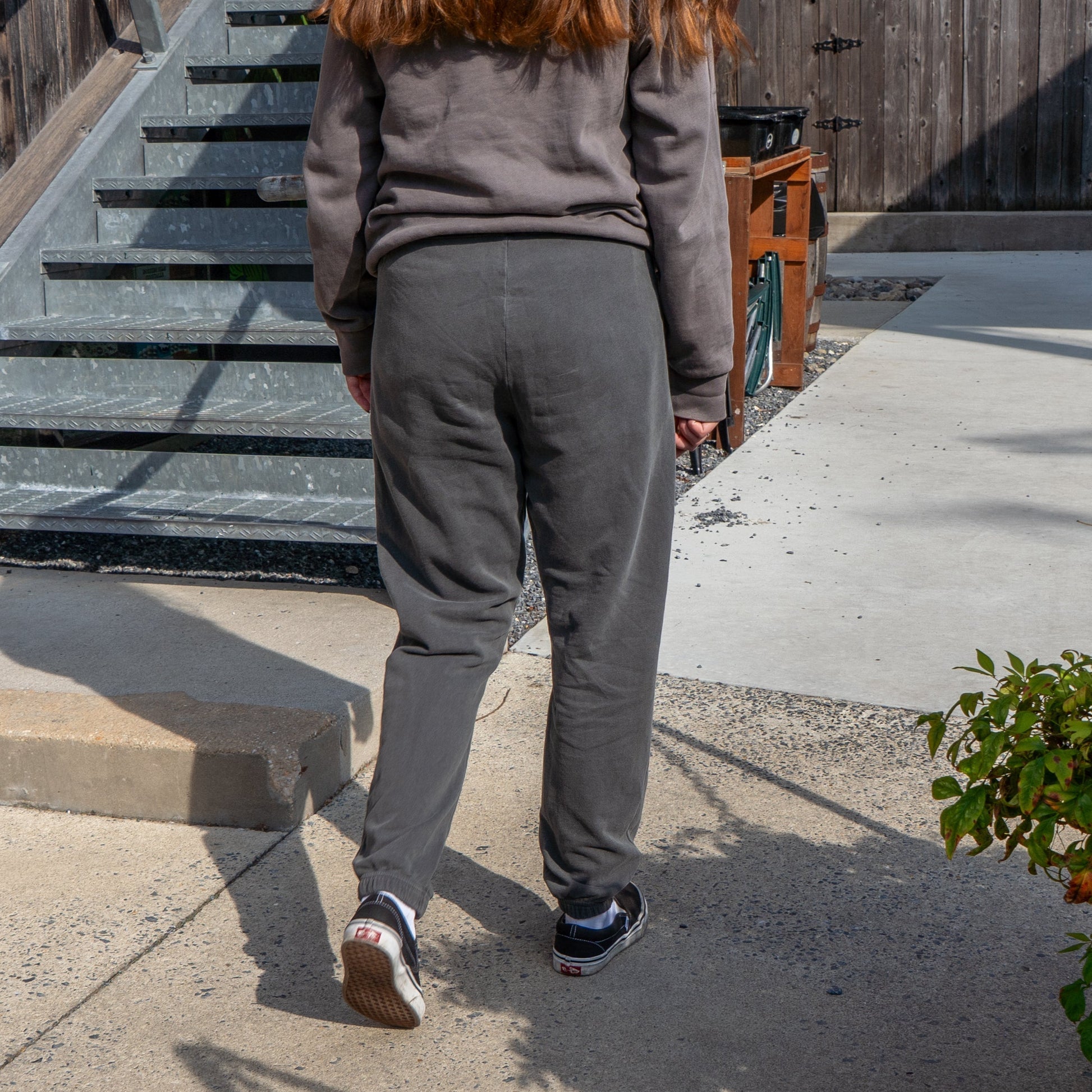 A person with long brown hair wearing GO WEST T-SHIRT COMPANY's Grey Got A Minute Sweatpants and a matching sweatshirt with black sneakers walks outdoors by stairs and a wooden fence as sunlight casts shadows on the ground.