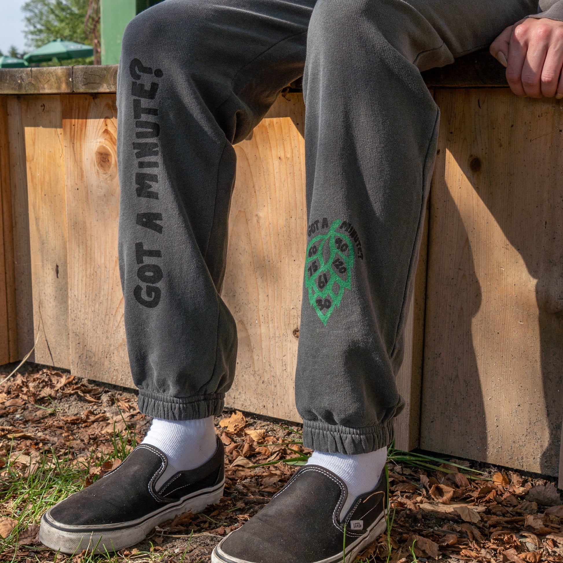 Relaxing outdoors on a wooden ledge, someone wears comfy Grey Got A Minute Sweatpants from GO WEST T-SHIRT COMPANY with "GOT A MINUTE?" on one leg, paired with white socks and black slip-ons among brown leaves and green grass.